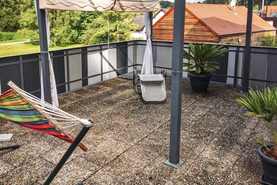 Takterrassen med gamla betongplattor erbjuder en rymlig vistelseyta. Den smutsiga ytan på betongplattorna visar tydliga tecken på slitage. Solskyddet ger behaglig skugga under varma dagar. Växtkrukor med palmer och en hängmatta skapar en inbjudande atmosfär. Rekkonstruktionen av metall och glas fullbordar terrassens moderna och öppna design.