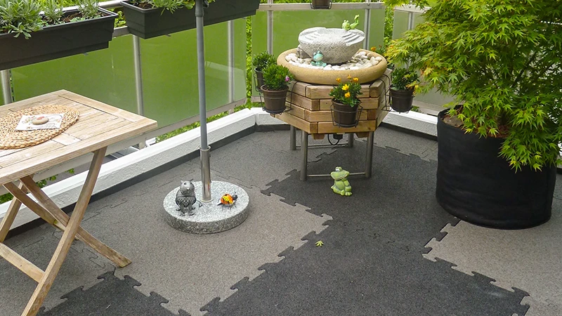 På bilden syns en inbjudande balkong belagd med tvåfärgade WARCO-pusselplattor. En liten fontän, en trähylla och flera blomlådor skapar en mysig atmosfär. Golvet ger hög komfort tack vare sin elastiska, halksäkra yta och är dessutom vädertåligt. Det exakta pusselsystemet möjliggör färgaccenter som ger utomhusmiljön en modern känsla.