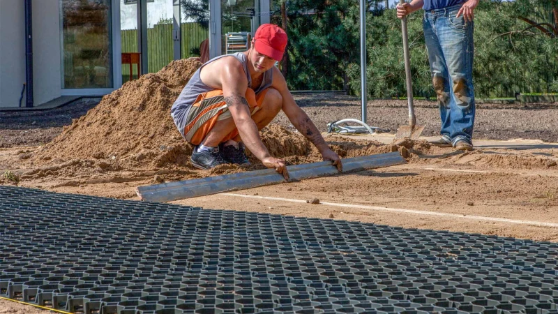Im Garten wird eine Tragschicht aus Kunststoffwabengitter verlegt. Die Wabengitter liegen auf einer Bettung aus Sand. Ein Mann ist gerade dabei, das Bett für die nächsten Bodengitter vorzubereiten und zieht den Sand über eine Lehre aus Rohren ab.