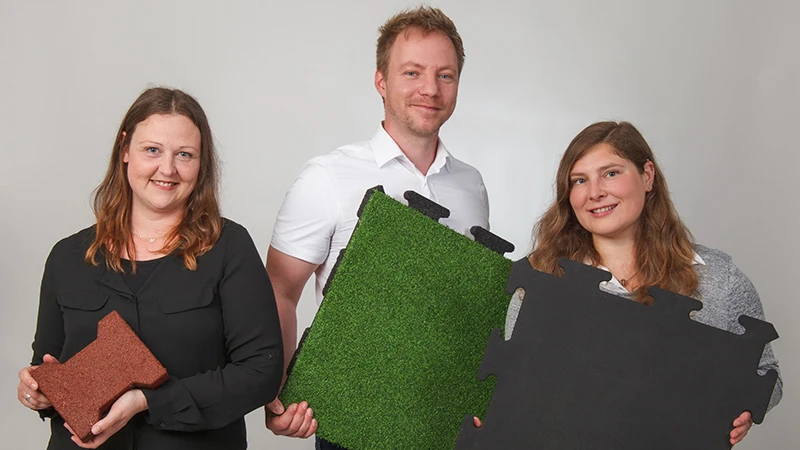 Med vänliga leenden visar tre WARCO-rådgivare upp golvplattor av PU-bundet gummigranulat i brunt, grönt och svart. Dessa plattor passar särskilt bra för balkonger och ger ett halksäkert, slitstarkt resultat. Varje yta har en egen design och är enkel att lägga. Tack vare deras expertkunskap hittar du rätt golvlösning för en säker och attraktiv balkong. De lättskötta plattorna erbjuder dessutom hög komfort.
