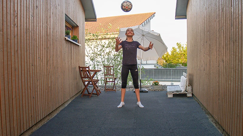 En flicka spelar boll på en terrass mellan två träklädda byggnadsväggar. Golvbeläggningen består av svarta terrassplattor av gummigranulat, vilket ger en robust och fjädrande yta lämplig för sportaktiviteter. I bakgrunden finns ett parasoll och en liten sittgrupp av trä som skapar en inbjudande atmosfär.