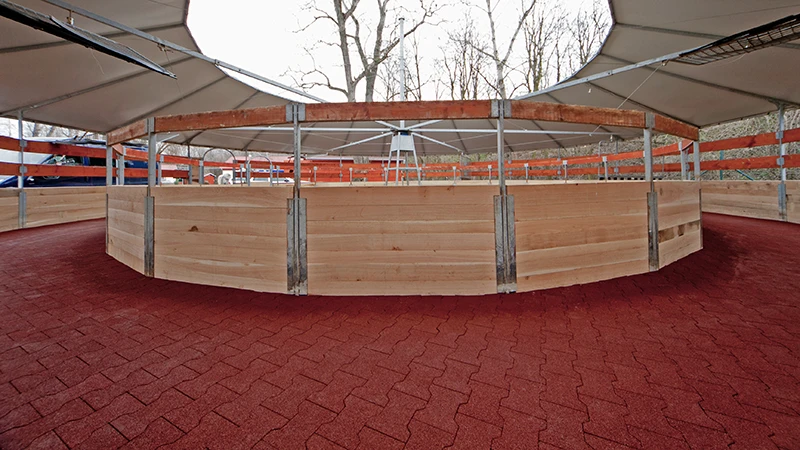 Bilden visar en modern, täckt hästwalker. Gångytan är helt belagd med röda gummiplattor – halksäkra, genomsläppliga och ledvänliga. Den elastiska ytan gör att hästarna kan röra sig säkert oavsett väder och gångart. Beläggningen är stabil, hållbar och mycket lätt att underhålla. Den passar perfekt för ridskolor, stuterier och hästkliniker som söker ett tyst, djuranpassat och slitstarkt underlag.