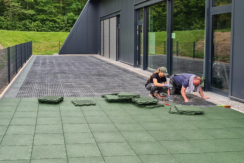 Mot en mörk, modern fasad installerar en kvinna och en man gröna WARCO-terrassplattor av PU-bundet gummigranulat. Plattorna läggs tätt för att skapa en halksäker, väderbeständig och hållbar yta på uteplatsen. Det robusta materialet ger komfort och skydd i alla väder. Den enkla installationen möjliggör personlig utformning och tillför varje uteplats en högkvalitativ, samtida karaktär. Plattornas tåliga yta bidrar dessutom till långvarig prestanda.