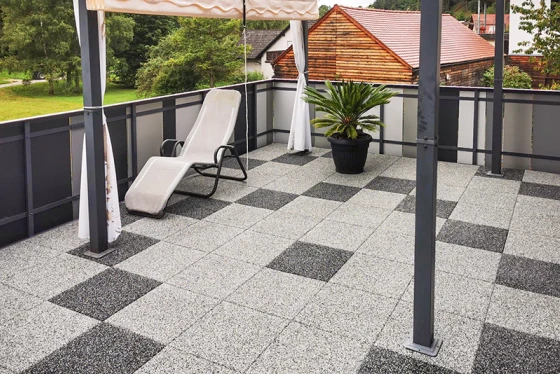 Den renoverade takterrassen är utformad med plattor i granitlook. De plattor i ett geometriskt mönster av ljusgrå och mörkgrå rutor skapar en modern och stilren atmosfär. Den robusta gummibeläggningen ger halkskydd och komfort. Ett solskydd ger behaglig skugga under varma dagar. En växtkruka med en palm och en solstol kompletterar den inbjudande terrassdesignen. Räckeskonstruktionen av metall och glas ger takterrassen en öppen och luftig känsla.