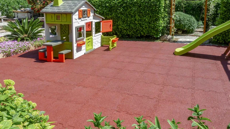 Spielplatz im Garten In der Mitte des Gartens, eingebettet in Sträucher, befindet sich eine Spielfläche aus ziegelrotem WARCO Spielplatzboden. Zum Spielen stehen den Kindern ein buntes Spielhaus und eine Kinderrutsche zur Verfügung.