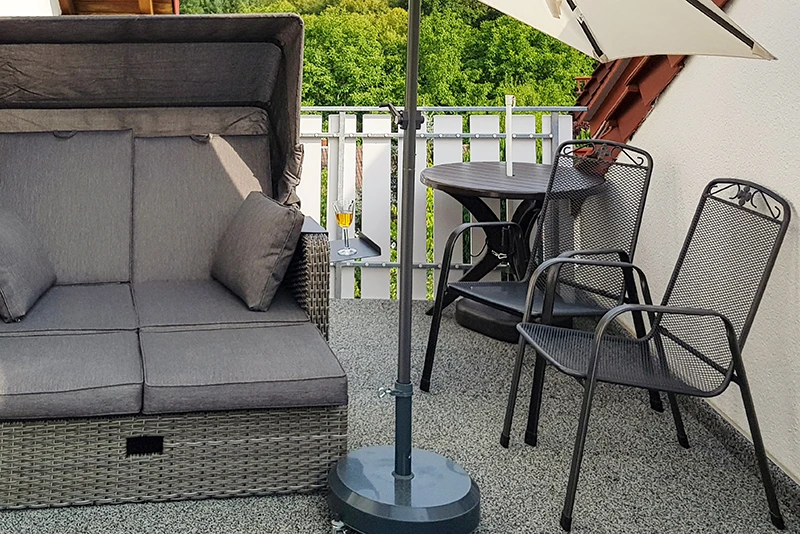 På bilden syns en balkong täckt av grå WARCO-golvplattor. En bekväm strandkorg och ett litet bord med två stolar i metall utgör en plats för avkoppling och njutning. Det elastiska, halksäkra golvet skyddar mot fukt och dämpar stegljud. Dessutom är det lättskött och vädertåligt, vilket gör att utrymmet förblir inbjudande och håller länge.