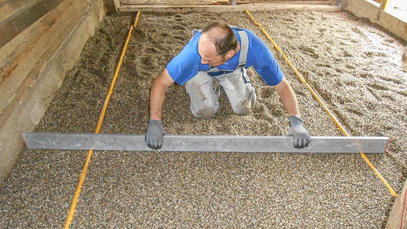 Installation av ett nytt golv i ett häststall: En man jämnar till ett gruslager med en rätskiva. Gruset fungerar som bädd för montering av ett gummibaserat boxgolv. Ytan begränsas av trä- eller betongväggar. Arbetet utförs noggrant och precist – typiskt för korrekt förberedelse av ett hästvänligt stallgolv.