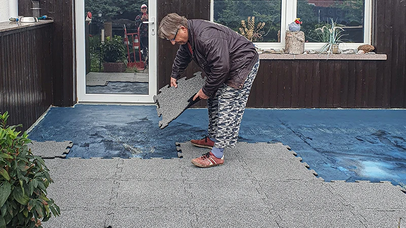 En terrass renoveras. En äldre kvinna lägger nästa 18 mm tjocka pusselplatta på den redan delvis täckta terrassen. Ett tätningsmembran fungerar som underlag för det nya golvbeläggningen av gummiplattor. Den exakta sammanlåsningen av plattorna möjliggör enkel och stabil installation.