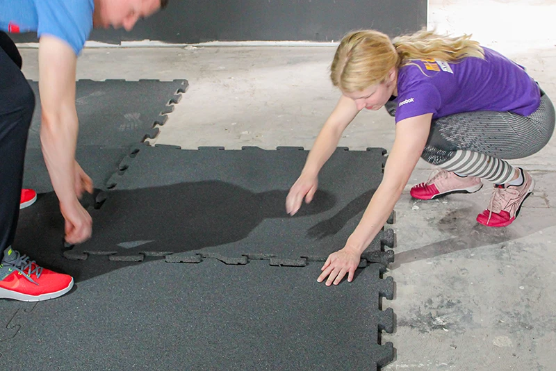 Zwei Personen verlegen graue Fallschutzplatten von WARCO auf einem Betonboden. Dank der Puzzle-Verzahnung lassen sich die Platten mühelos und passgenau miteinander verbinden. Die einfache Handhabung ermöglicht eine schnelle Installation, ohne den Einsatz von Werkzeugen. Diese stoßdämpfenden Fallschutzplatten sind ideal für Bereiche wie Fitnessstudios, Spielplätze oder andere Räume, die einen sicheren und langlebigen Bodenbelag erfordern. WARCOs Fallschutzplatten bieten eine hervorragende Kombination aus Sicherheit, Schutz vor Verletzungen und einfacher Montage.